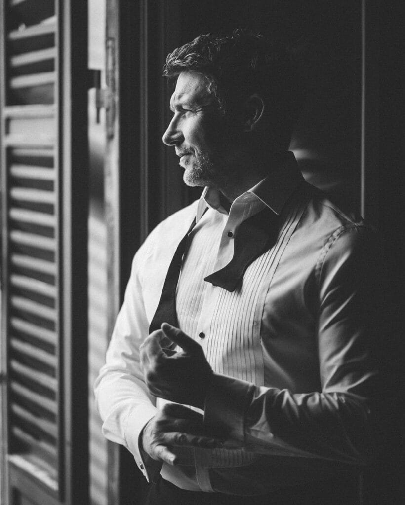 A man in a formal white shirt with a loose bow tie stands by a window at Villa Palazzola. The black and white photo captures his thoughtful expression, as light and shadows create patterns on the wall, embodying the artistry of a wedding photographer in Italy.