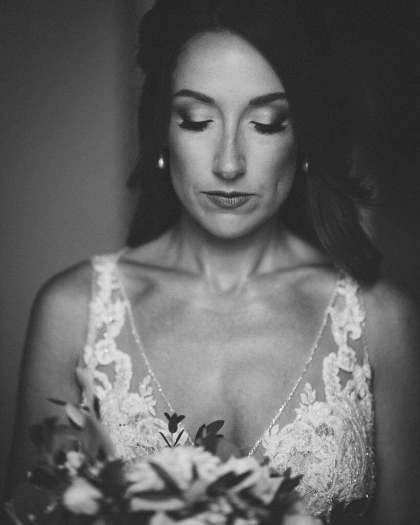 Black and white photo of a bride in a lace wedding dress at Villa Palazzola. She is looking down at a bouquet with a calm expression, captured beautifully by an expert Rome wedding photographer in Italy. The image highlights intricate dress details and the soft texture of the flowers.