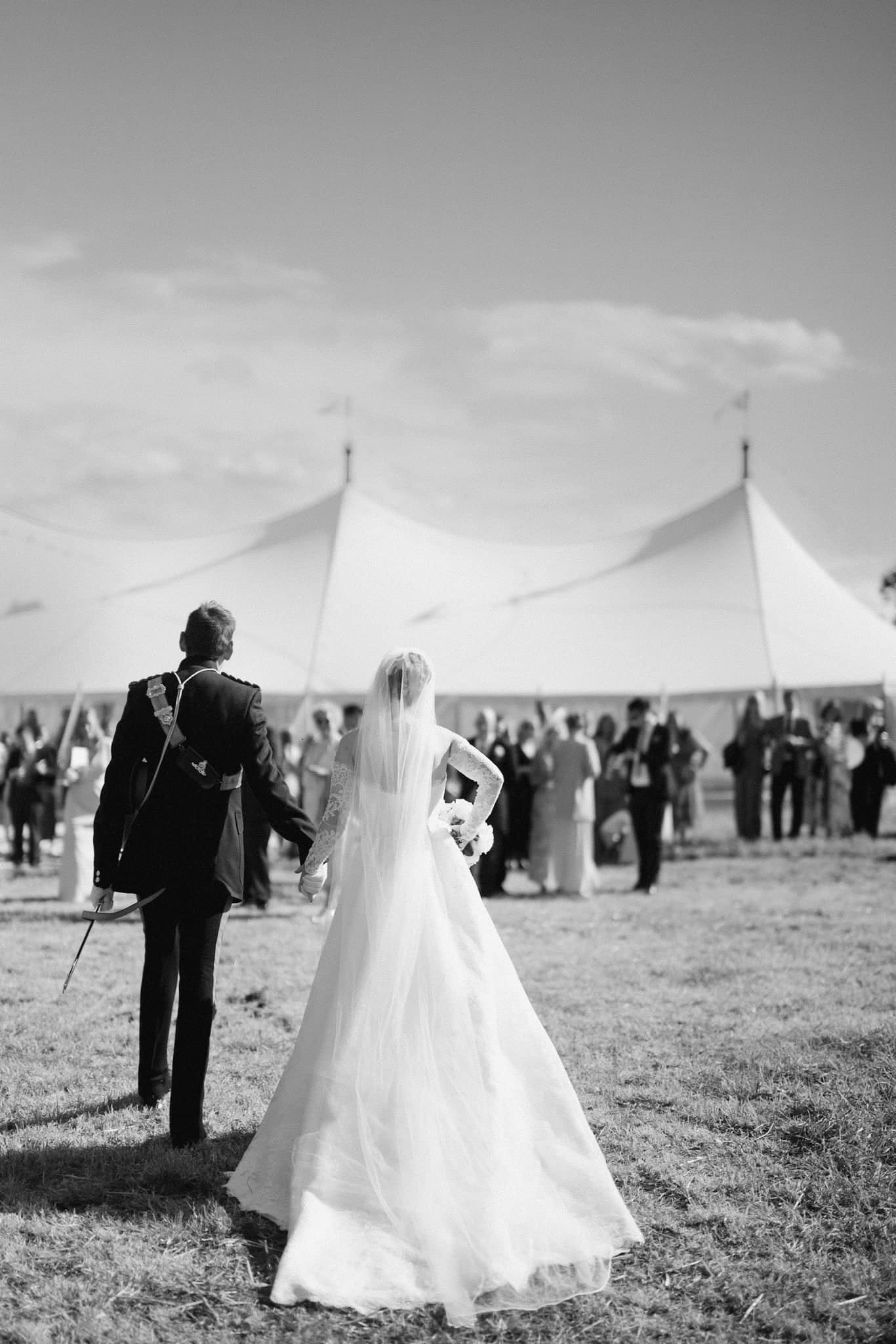 A bride and groom walk hand in hand towards a large white marquee tent, embodying the ultimate guide to marquee garden wedding receptions. The bride’s long, flowing gown and veil complement the grooms dark suit. Guests gather near the tent in an open, grassy area under a clear sky. Black and white photo.