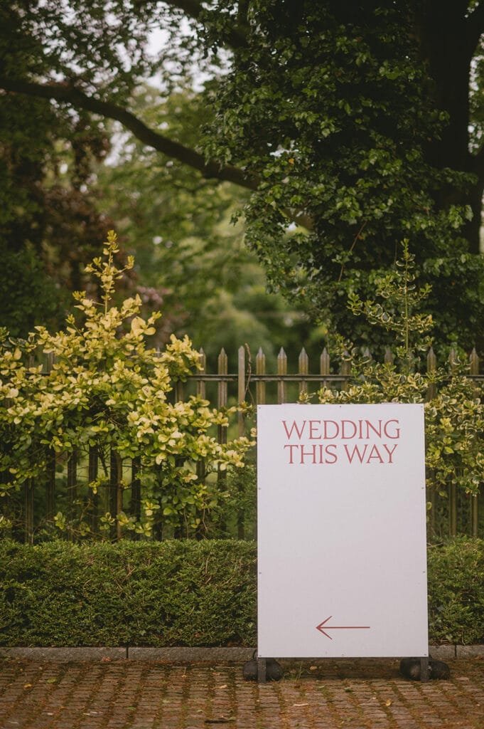 In a charming garden setting with tall trees and a wooden fence in the background, a rustic sign directs guests: Wedding This Way. Ideal for capturing stunning shots, its a perfect spot for a creative wedding photographer to capture those cherished moments.