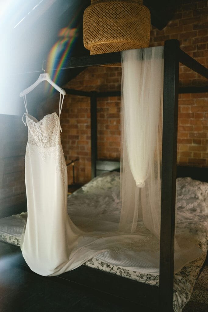 A white lace wedding dress hangs elegantly on a black canopy bed in a rustic room with brick walls, captured by a talented pumping house wedding photographer. Sunlight casts a slight rainbow effect, and a long veil drapes over the bed beneath a woven lampshade above.