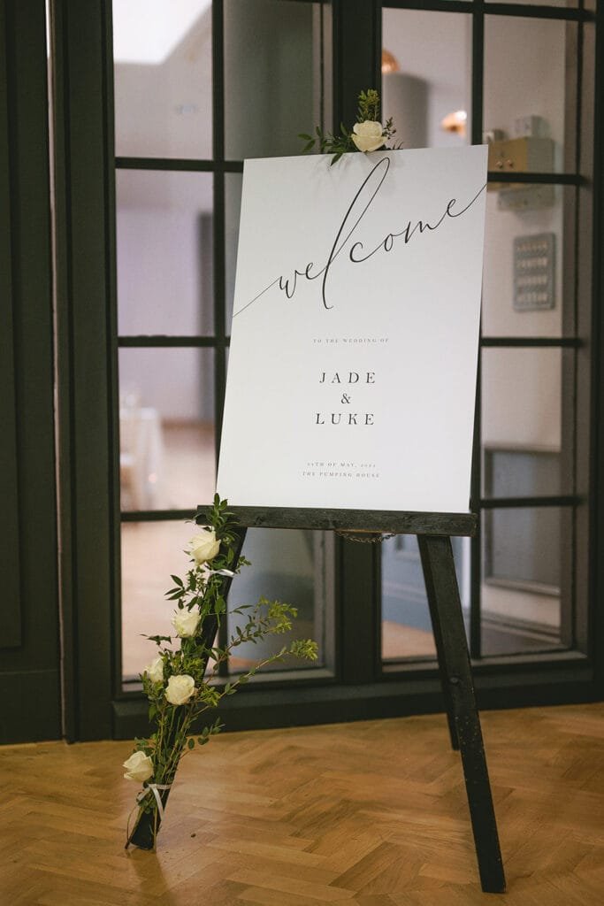 A wedding welcome sign on an easel, captured gracefully by a pumping house wedding photographer, features Jade & Luke in elegant script. Decorated with small white flowers and greenery, it stands against dark framed glass doors and light wooden flooring.