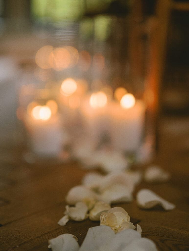 Soft focus image of lit candles on a wooden surface, surrounded by scattered white rose petals. The warm candlelight creates a cozy and intimate atmosphere with a blurred background, capturing the essence of a pumping house wedding as any skilled photographer aims to achieve.