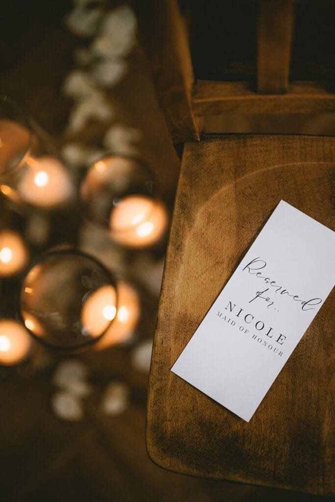 A wooden chair with a white card that reads Reserved for Nicole, Maid of Honour. Nearby, soft candlelight reflects in glass holders with rose petals scattered on the floor. Captured by a wedding photographer at the pumping house, the scene creates a warm and intimate atmosphere.