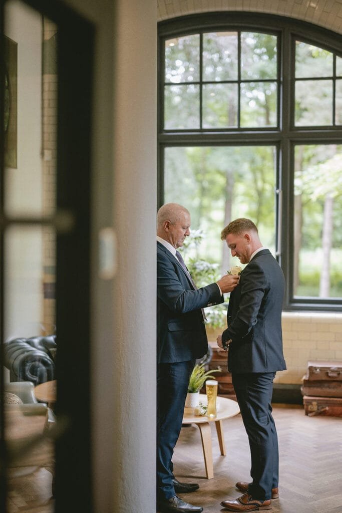 Two men in suits stand by a large window in an elegantly decorated room, preparing for the big day. One man is adjusting the others tie. The room features leather chairs, a round wooden table, and large suitcases as decor. A pumping house wedding photographer captures it all with trees visible through the window.