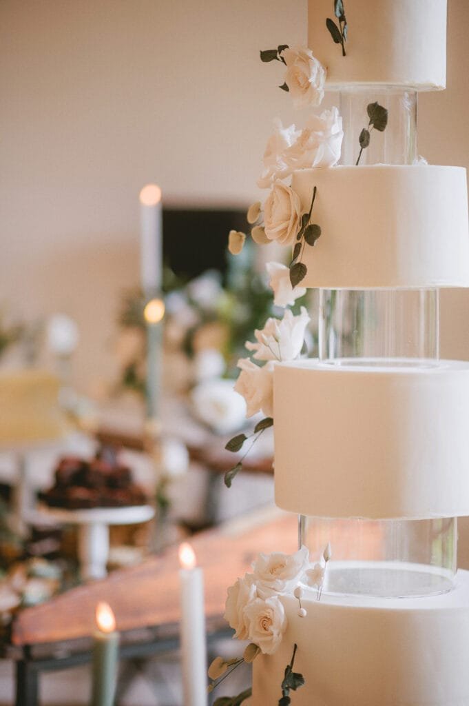 A tall, elegant wedding cake with multiple tiers, each separated by clear acrylic pillars. Captured by a skilled pumping house wedding photographer, the cake is adorned with white roses and green leaves, against a blurred backdrop of a table with candles and flowers.