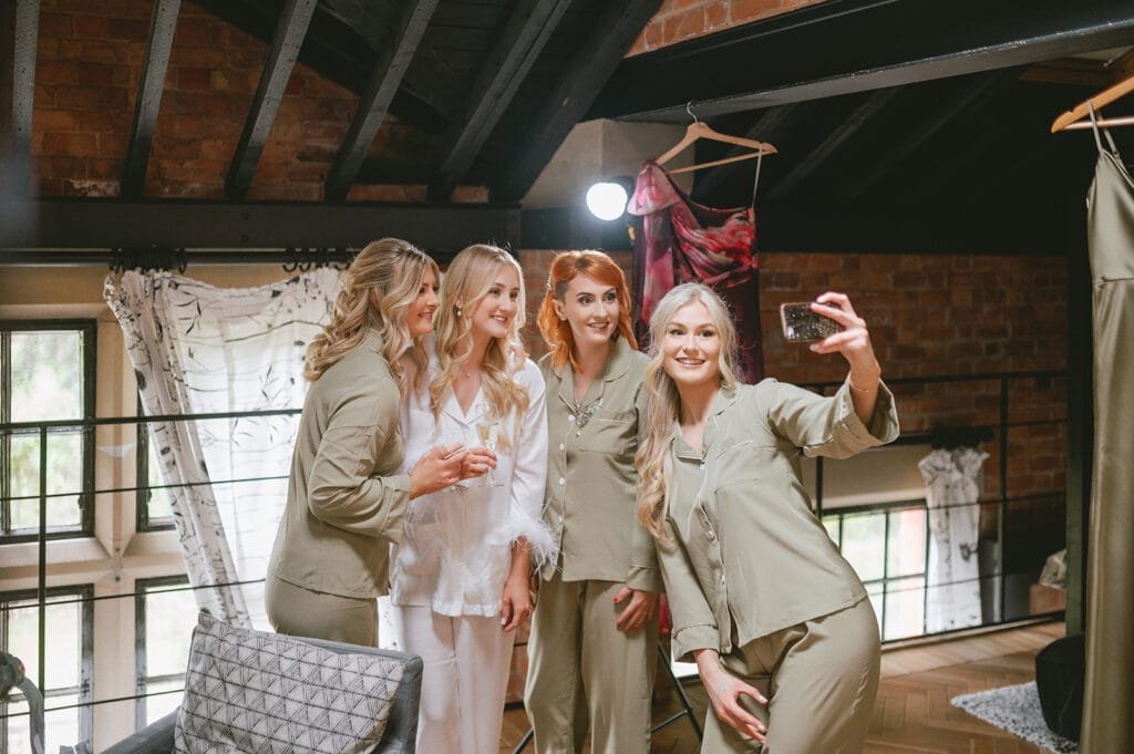 Four women in pajamas take a selfie in a cozy room. Three wear matching olive pajamas, and one wears a white set. With smiles that could light up any wedding photo, they stand close together against brick walls and decorative fabrics.