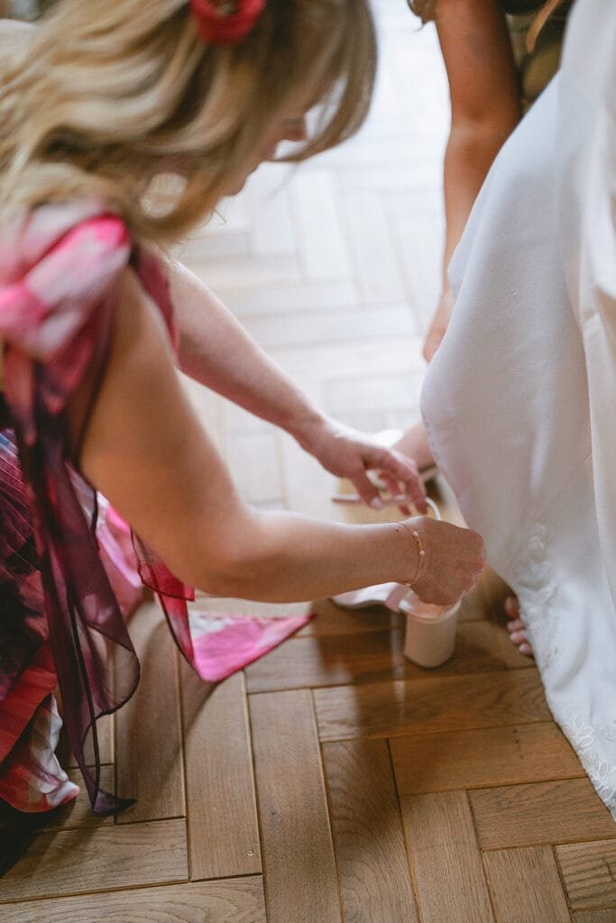 A woman in a colorful dress kneels on a wooden floor, assisting with the adjustment of a white high-heeled shoe for another person in a white dress. Captured by the wedding photographer, the scene suggests preparation for an event, possibly set in the charming ambiance of a pumping house.