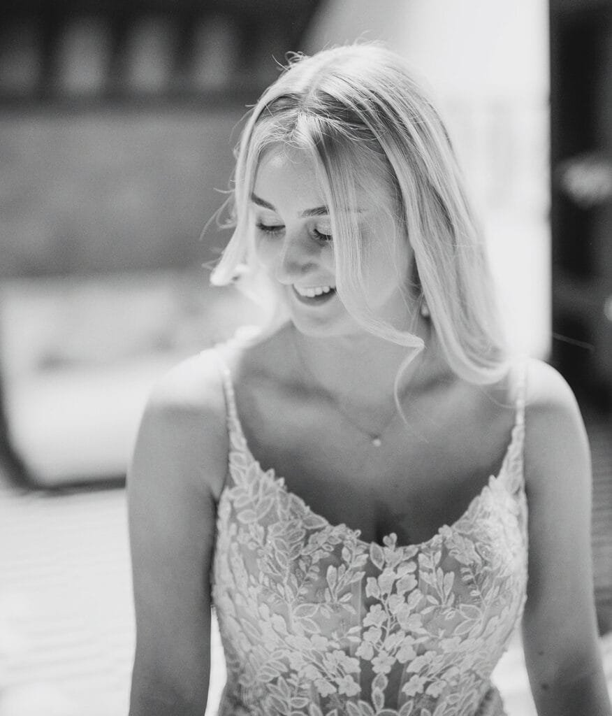 A black and white photo of a smiling woman in a lace dress with floral patterns, perhaps captured by a talented wedding photographer. Her long hair falls softly around her face as she gazes downward. The background is blurred, emphasizing her as the focal point.