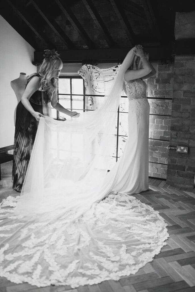 A bride stands in a loft-style room, adjusting her veil. Her long, flowing train is being arranged by her mumt. The room, perfect for a pumping house wedding photographer, features exposed brick walls, a mannequin, and wooden flooring. Black and white photo.