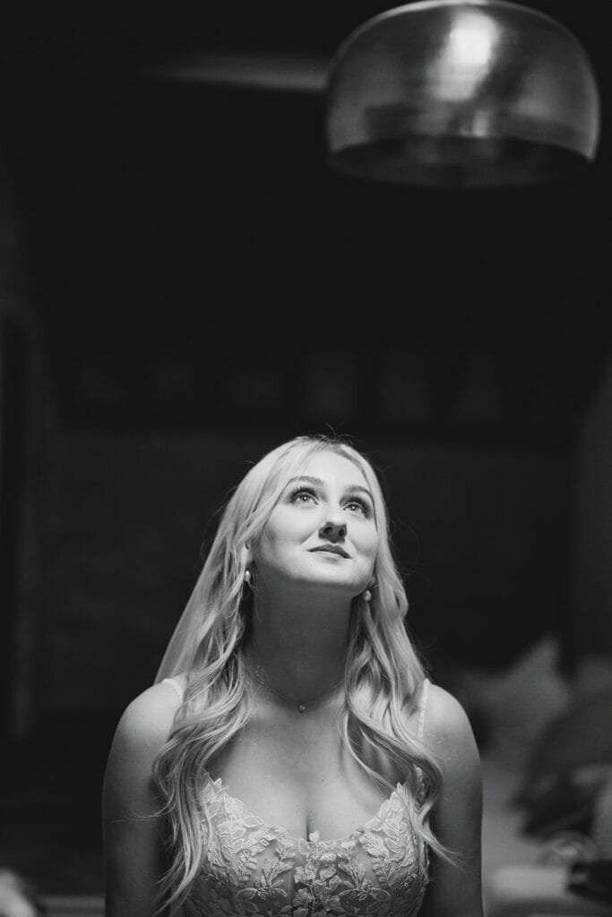 A bride in a detailed lace wedding dress gazes upwards with a serene expression, captured by a skilled wedding photographer. Her long, wavy hair cascades over her shoulders beneath the soft lighting of the circular fixture above. This elegant, black and white image conveys timeless beauty.