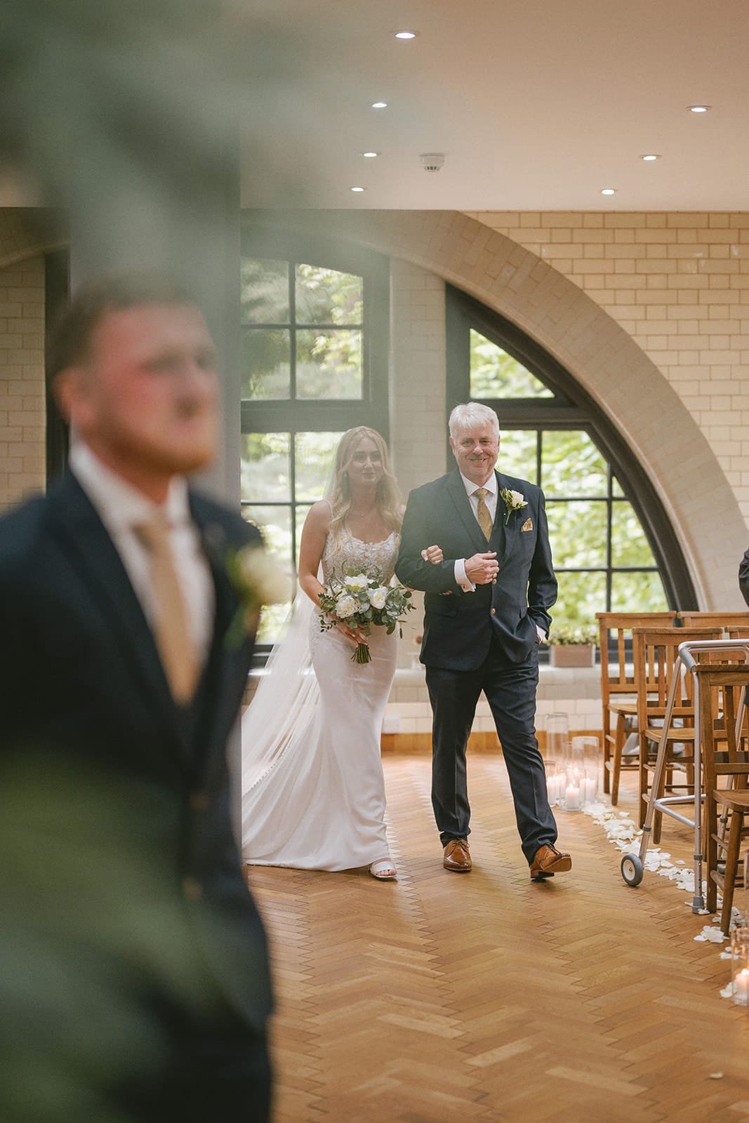 A bride in a white gown gracefully walks down the aisle, escorted by an older man in a suit. In the foreground, a pumping house wedding photographer captures every precious moment. Large arched windows and rows of chairs frame this beautiful scene.