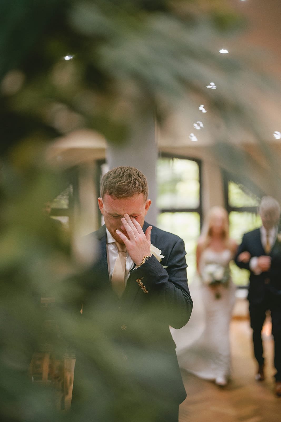 A groom stands with his hand covering his face, perhaps emotional or anxious, in a softly lit room captured by the wedding photographer. In the background, a bride holding a bouquet walks down the aisle towards him, accompanied by an older man. The scene is beautifully framed with blurred greenery.