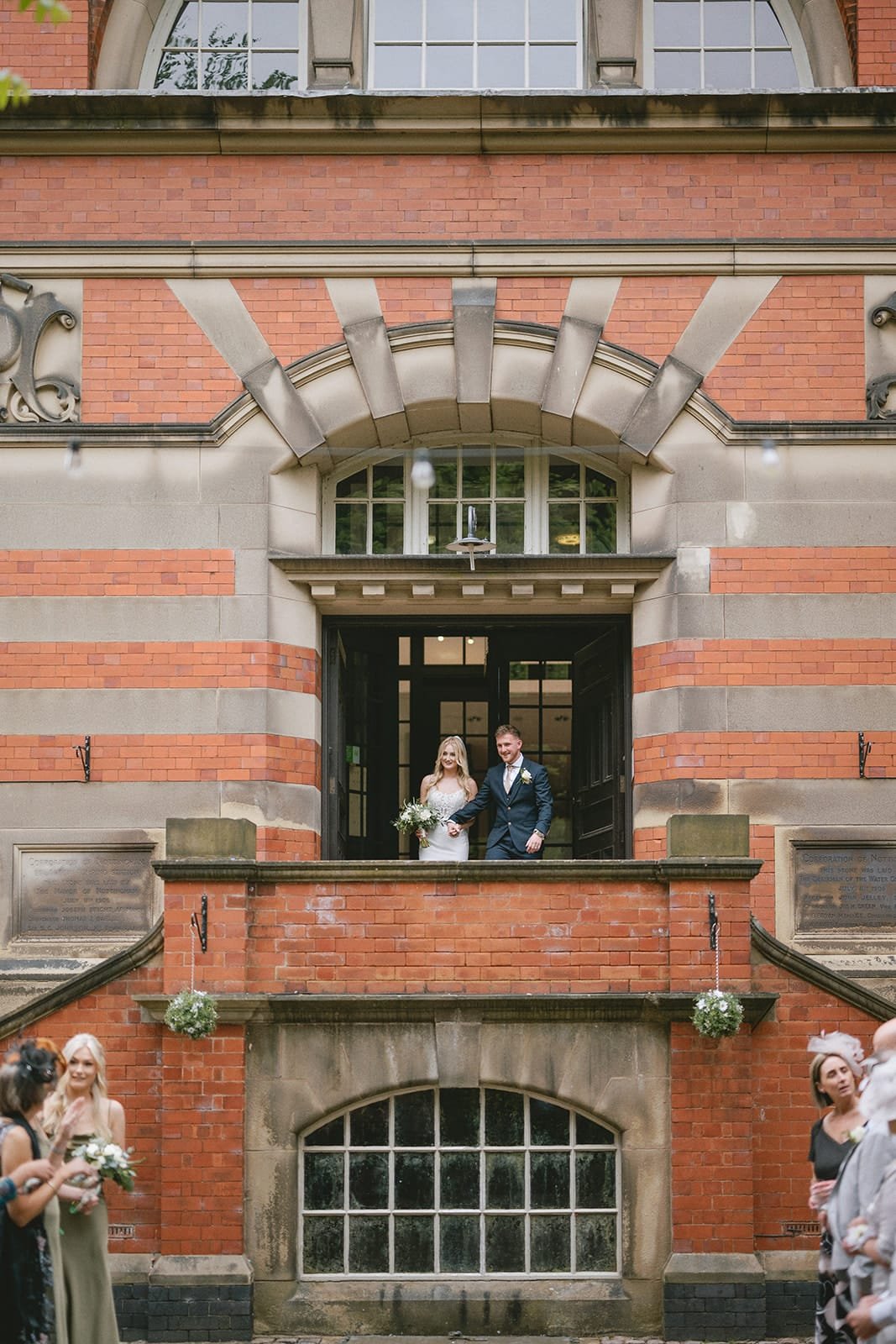 The bride and groom stand on a balcony of the charming red brick building, elegantly captured by their pumping house wedding photographer. Guests gather below as the bride clutches her bouquet, both smiling in formal attire amidst large arched windows and intricate stonework.