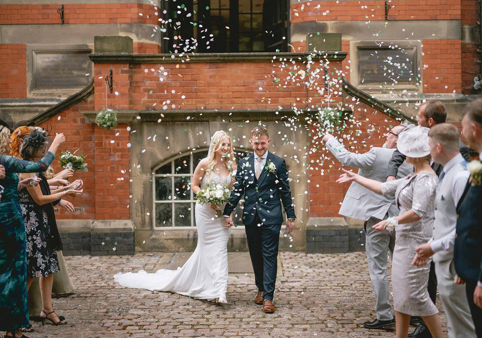 A newlywed couple walks hand in hand, smiling as confetti fills the air, captured perfectly by a skillful wedding photographer. The bride, in her white gown with flowers, and the groom, in his dark suit, stand on a cobblestone path before a charming brick pumping house surrounded by cheering attendees.