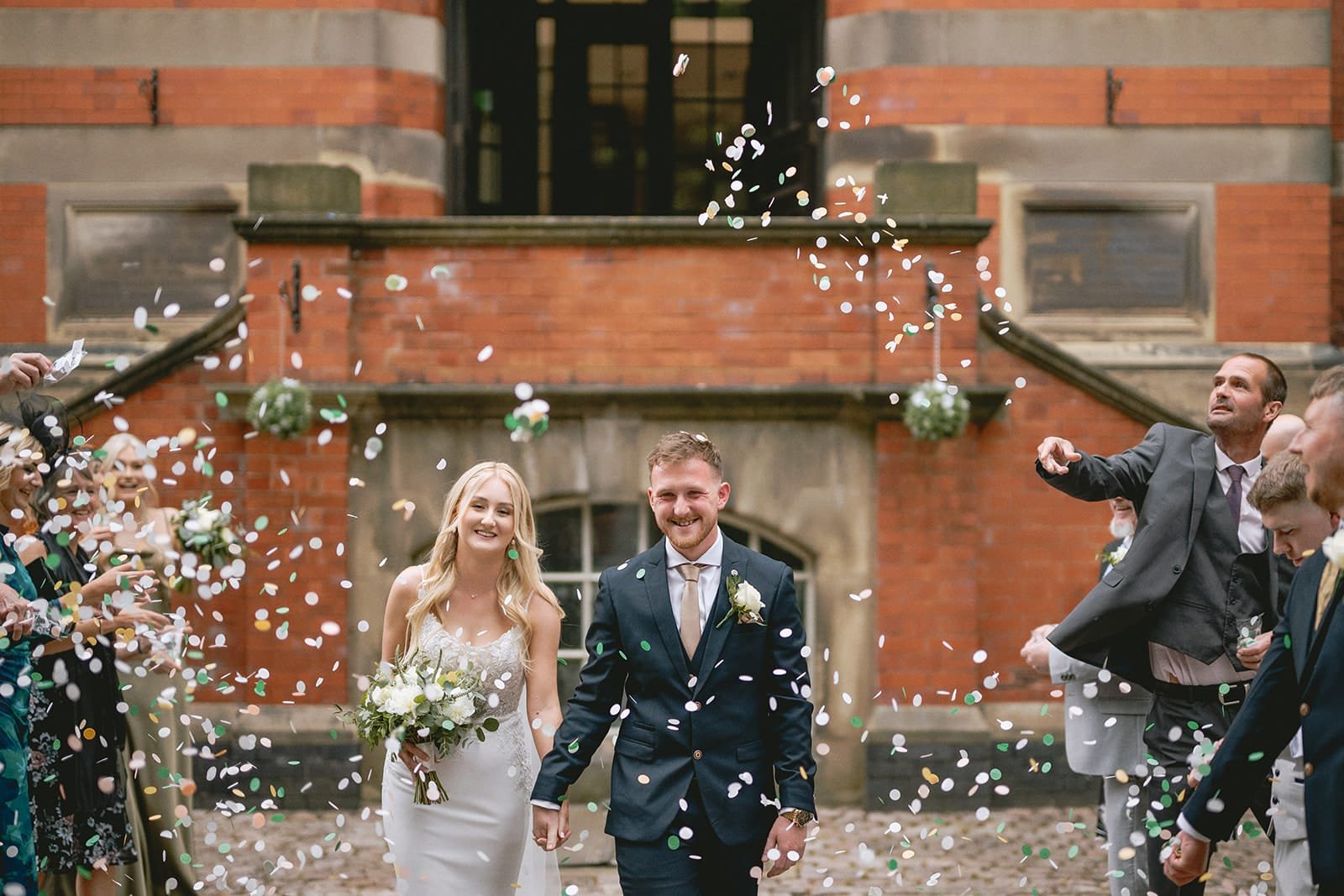 A bride and groom walk hand in hand outside the historic pumping house, smiling as guests throw confetti over them. The bride holds a bouquet of white flowers, and the groom wears a dark suit. A joyful crowd surrounds them, celebrating their union while a wedding photographer captures every cherished moment.