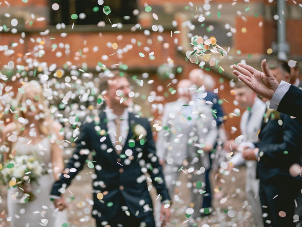 Confetti fills the air as a bride and groom, surrounded by guests, celebrate outdoors. The couple holds hands, the groom is in a dark suit. A pumping house wedding photographer captures this festive scene, with blurred guests watching and celebrating.