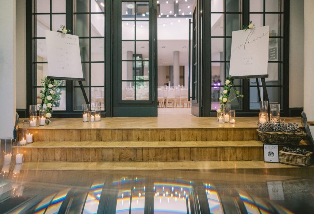 Elegant entrance with double glass doors, two easels bearing white signs, floral decorations, and lit candles. Steps lead to a stylish indoor venue, perfect for capturing moments as a pumping house wedding photographer, with a wicker basket and small blackboard on the side.