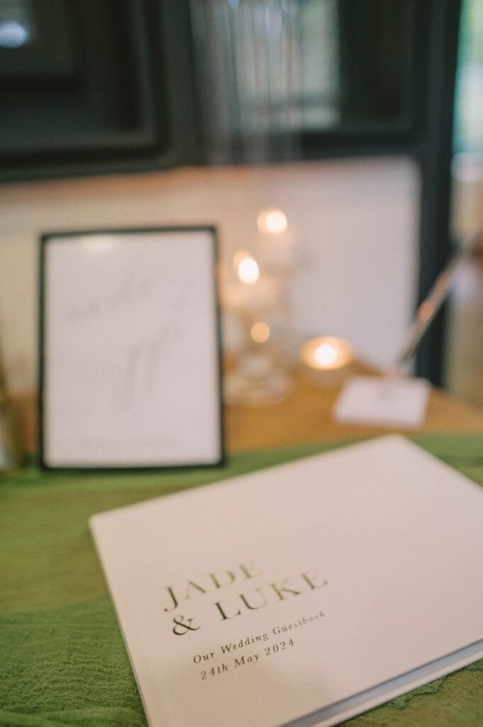 A wedding guestbook titled Jade & Luke: Our Wedding Guestbook, 24th May 2024 lies open on a table. In the background, captured by a talented pumping house wedding photographer, a frame and several lit candles create a warm ambiance.