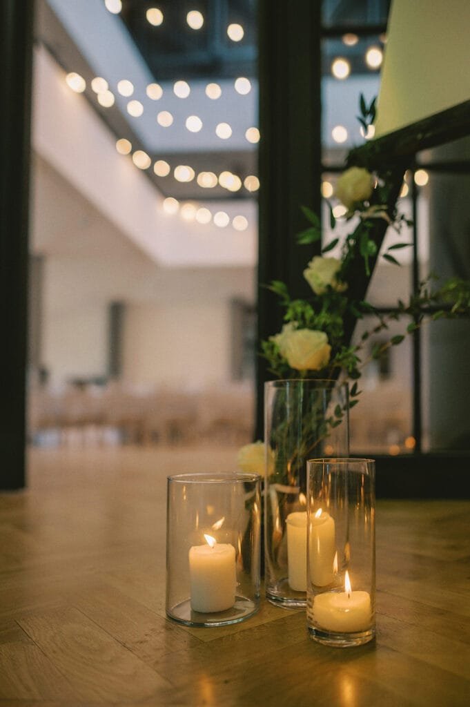 Three lit candles in glass holders rest on a wooden floor, casting a warm glow. Behind them, a tall vase holds white roses and green foliage. The softly lit room, ideal for a pumping house wedding photographer, features blurred string lights above.