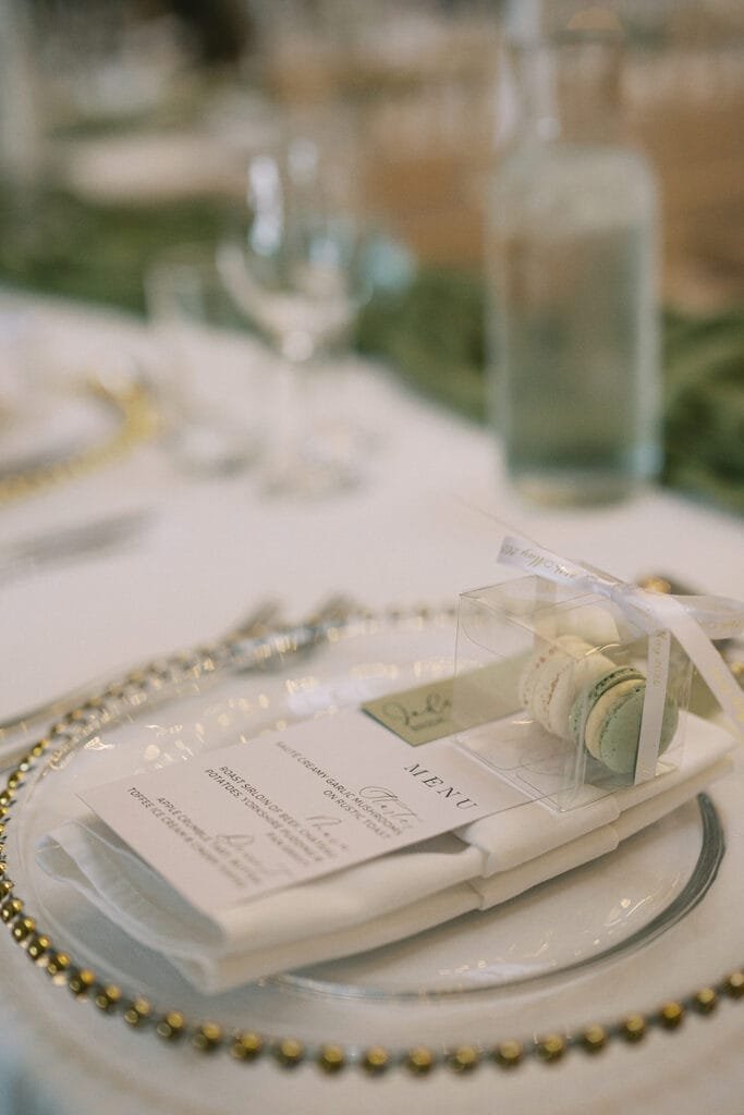 Elegant wedding table setting at a Pumping House celebration, with a decorative plate featuring a gold beaded rim. On it rests a folded menu and a clear box containing three macarons tied with ribbon. A water bottle and glasses are visible in the blurred background, ready for capture by the photographer.