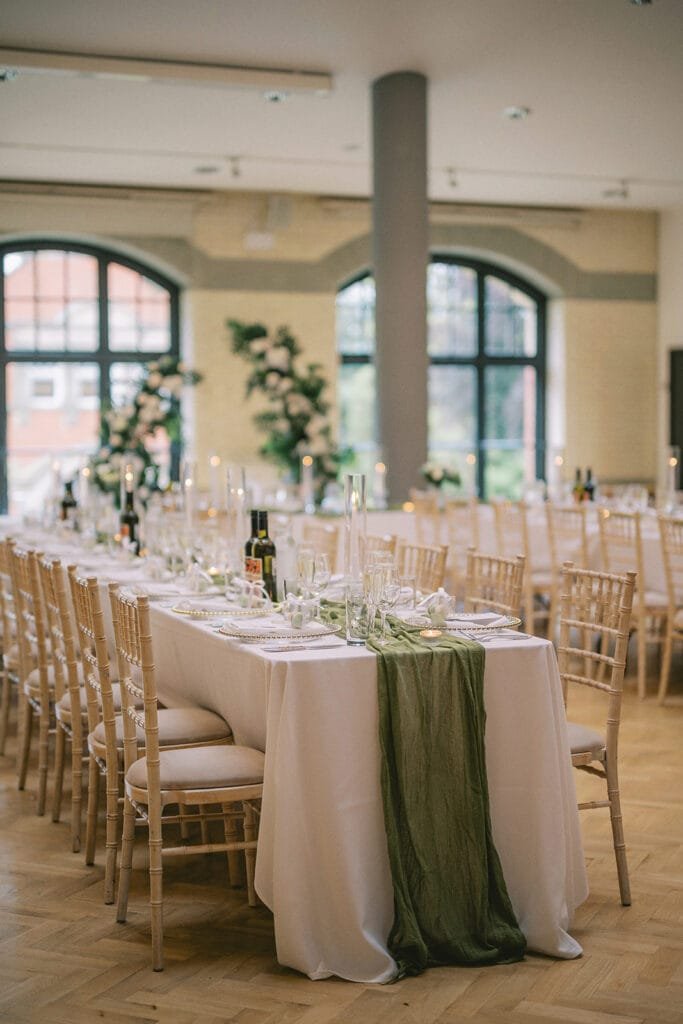 Elegant wedding reception setup with long tables draped in white cloths and green runners, captured beautifully by a pumping house wedding photographer. Each table is adorned with candles, glassware, and greenery. The venue boasts high ceilings, large arched windows, and wooden chairs.