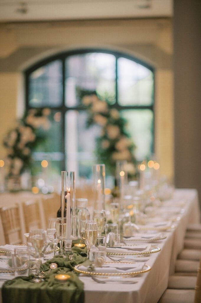 Elegant wedding reception table set with white linens, gold-rimmed plates, crystal glasses, and tall candles. Green foliage and flowers decorate the center. Soft lighting and a large arched window in the background create a warm atmosphere perfect for a Pumping House wedding photographers lens.