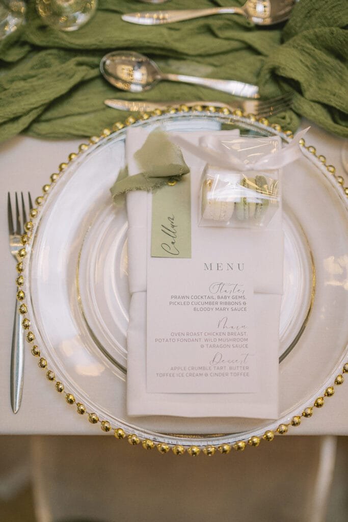 A beautifully set table captures the charm of a pumping house wedding. At its center, a clear plate with a decorative gold beaded rim cradles a green fabric napkin beneath the menu and wedding favor. Silverware surrounds it, while dishes like prawn cocktail and chicken await their moment.