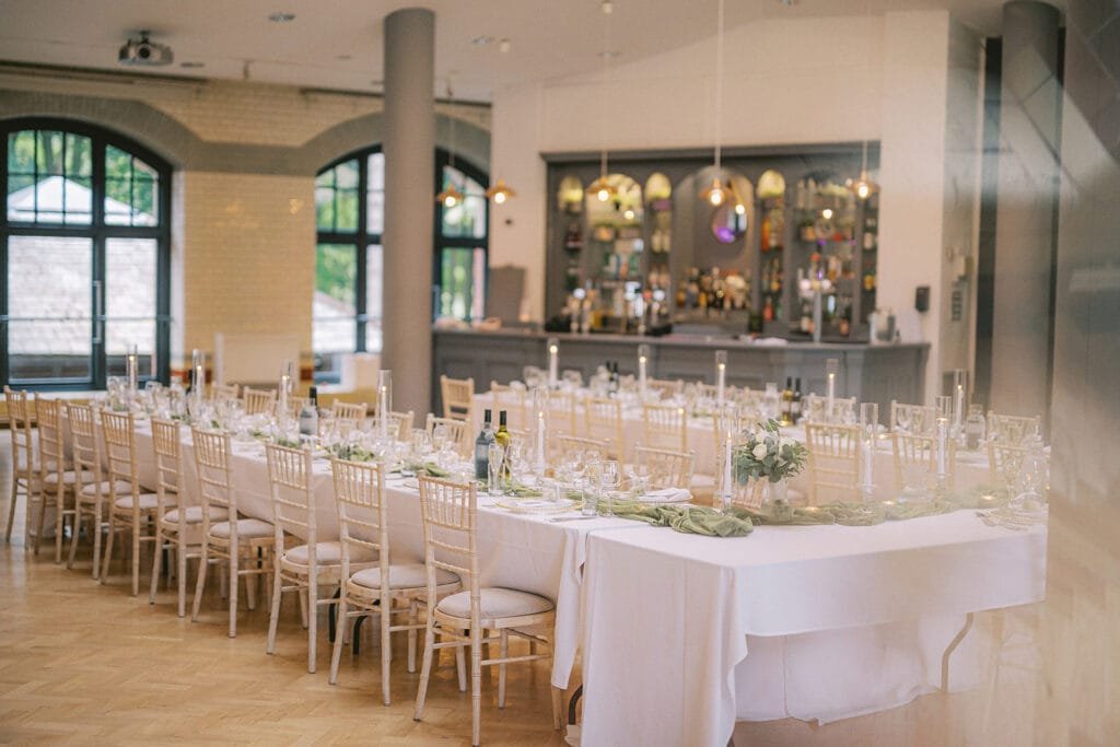 A spacious banquet room with elegantly set tables arranged in a U-shape awaits, perfect for capturing by a wedding photographer. Tables draped in white cloths feature green table runners, candles, and floral centerpieces. A bar gleams in the background as large windows invite natural light inside.