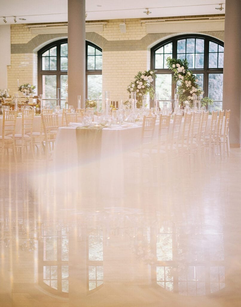 Elegant banquet hall with long tables set for a formal event, perfect for capturing by a wedding photographer. Tables adorned with tall floral arrangements and candles. Large arched windows grace the background, while the floors reflection mirrors the setup, creating a dreamy effect.