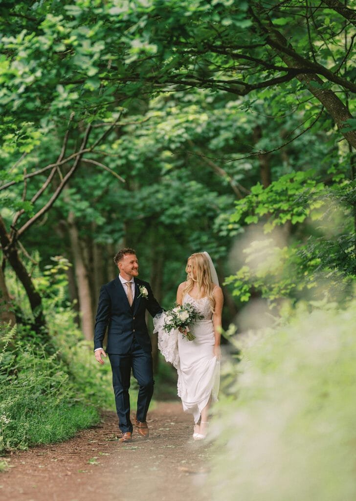 A bride and groom walk hand in hand along a forest path. The groom is in a navy suit, the bride wears a white dress and holds a bouquet. Lush green foliage surrounds them, creating a serene, romantic setting captured beautifully by the pumping house wedding photographer.