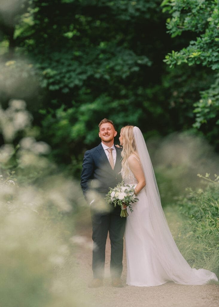 A bride and groom stand on a path surrounded by lush greenery. The bride, in a flowing white gown with veil, holds a bouquet and gently kisses the grooms cheek. Captured by a talented wedding photographer, the groom in a dark suit smiles blissfully with eyes closed.