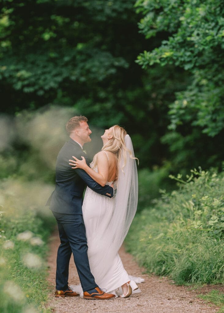 A bride in a white dress and veil and a groom in a dark suit share a joyful moment on a lush, green forest path. Captured by their talented wedding photographer, they laugh together, surrounded by tall trees and greenery.