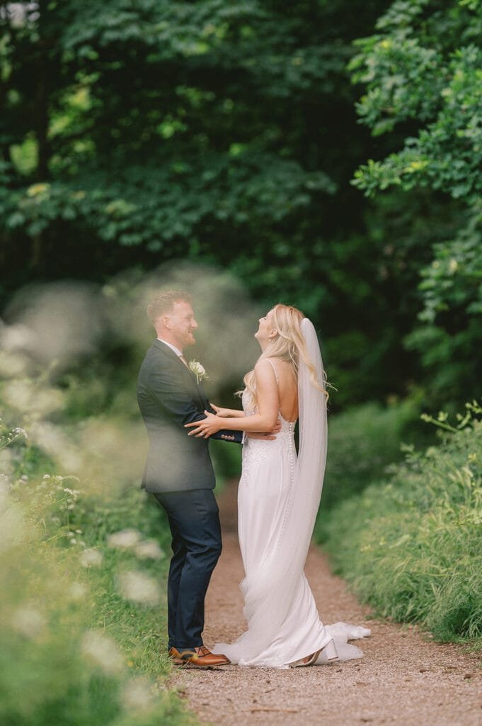 A bride and groom stand on a pathway in a lush, green forest setting. The bride wears a white gown with a long veil, and the groom is in a dark suit with a boutonniere. Captured by their talented wedding photographer, they hold hands, smiling at each other amidst nature’s charm.