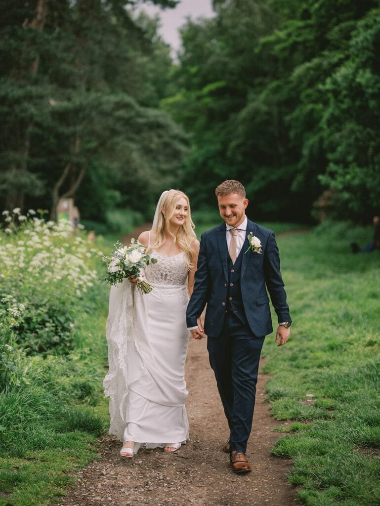 A bride and groom walk hand in hand through a lush, green park, captured beautifully by their wedding photographer. The bride dazzles in a white dress with a bouquet, while the groom is sharp in a dark suit. Both are smiling and gazing at each other lovingly.