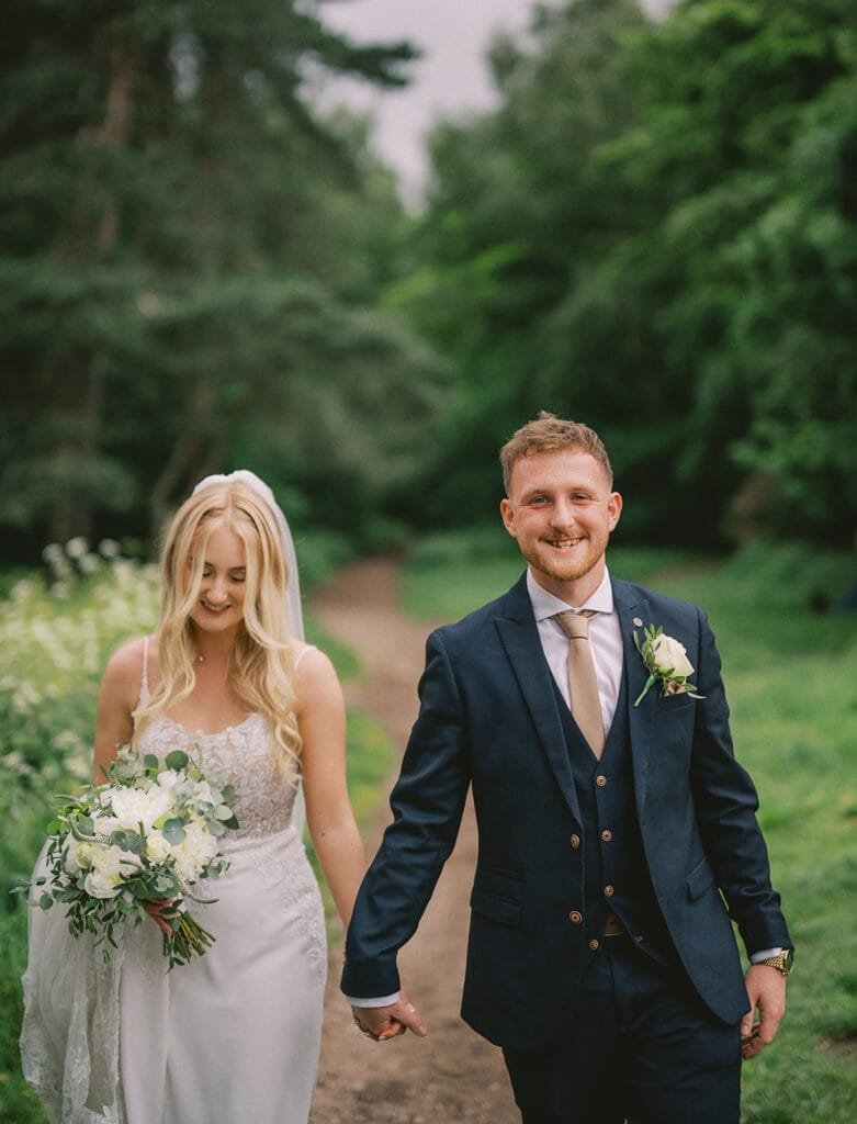 A couple strolls hand in hand along a tree-lined path, captured perfectly by their wedding photographer. The woman, in a flowing white dress with a bouquet of delicate blooms, and the man in his navy suit with a boutonniere, both smile amidst the lush greenery near the charming pumping house venue.