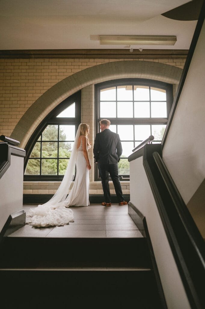 A bride and groom stand together at the top of a staircase, gazing out a large window. The scene, captured by a talented wedding photographer, features the bride in a long white gown and the groom in a dark suit, set against the pumping houses arched windows and brick walls.