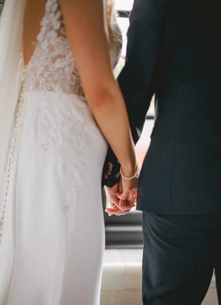 A bride and groom stand side by side, holding hands. The bride wears a white lace wedding dress, and the groom is in a dark suit. Their backs are partially turned, capturing an intimate moment that any skilled wedding photographer would treasure at a charming pumping house venue.