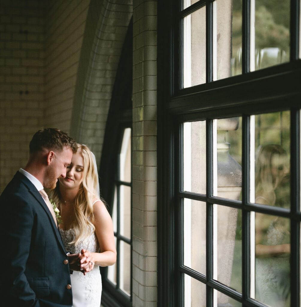 A couple stands close together near a large window, the architecture like an elegant backdrop. The wedding photographer captures the man in a dark suit gently holding the womans hand, her white dress shimmering softly. Both appear serene, gazing downward with soft smiles.pumping house wedding photographer