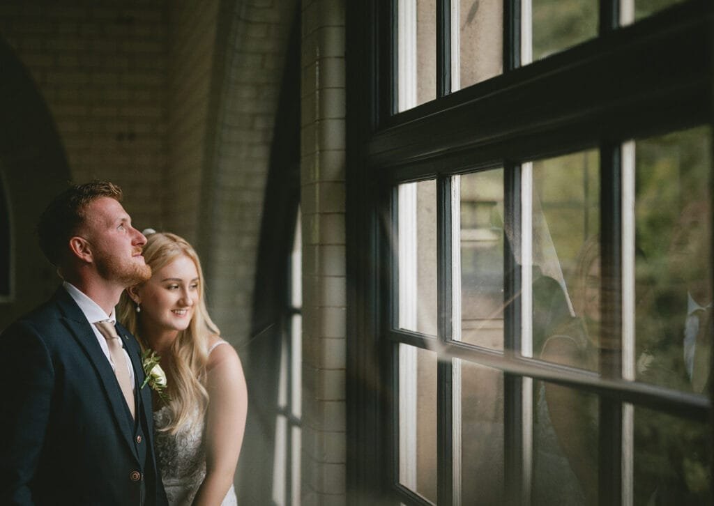 A couple dressed in wedding attire stands together, gazing serenely out of a large window. The groom, in a dark suit with a boutonniere, and the bride, with long blonde hair and a white dress, are captured beautifully by their pumping house wedding photographer as light streams through.