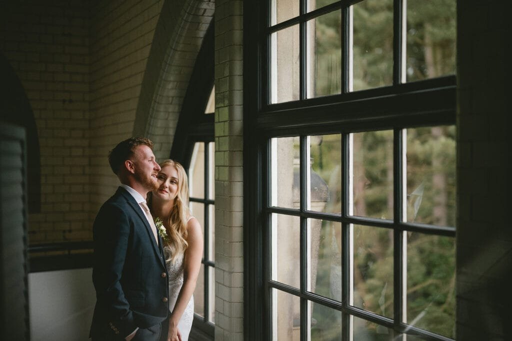 A couple stands indoors near large windows, captured by a talented wedding photographer. The man, in a dark suit, looks left as the woman, in a light dress, smiles gently at him. Through the windows of this charming pumping house venue, lush greenery creates a soft, natural backdrop.