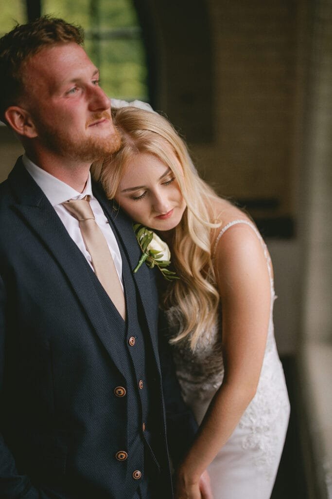 A bride in a white lace gown leans her head on a grooms shoulder. He wears a dark suit with a beige tie and boutonniere. Captured by a skilled wedding photographer, they stand indoors at the pumping house, soft light illuminating their serene, intimate atmosphere.