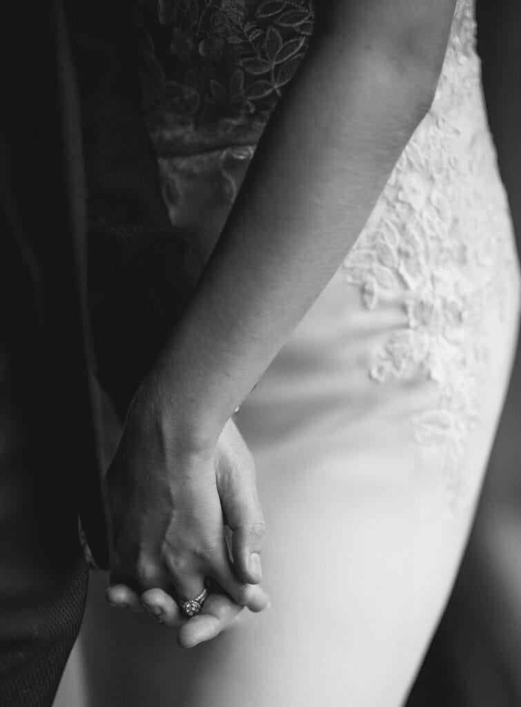 Black and white image of a couple holding hands, captured by a talented wedding photographer. The focus is on the hands, one adorned with a ring. The person on the right wears an elegant lace dress while the other dons a sharp dark suit.