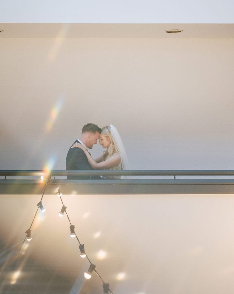 A bride and groom stand closely on a balcony, embracing each other. The gentle sunlight creates a dreamlike atmosphere with lens flares, beautifully captured by the Pumping House wedding photographer. A string of lights is visible in the foreground, adding to the charm.