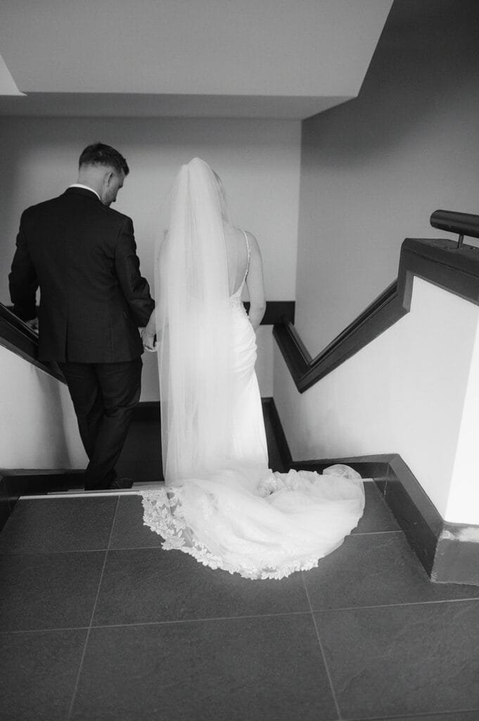 A black and white photo captures the bride and groom, hand in hand, descending a staircase. Her gown flows elegantly alongside her veil, while he is sharp-suited. This timeless moment, artfully framed by the pumping house wedding photographer, showcases their journey together from behind.