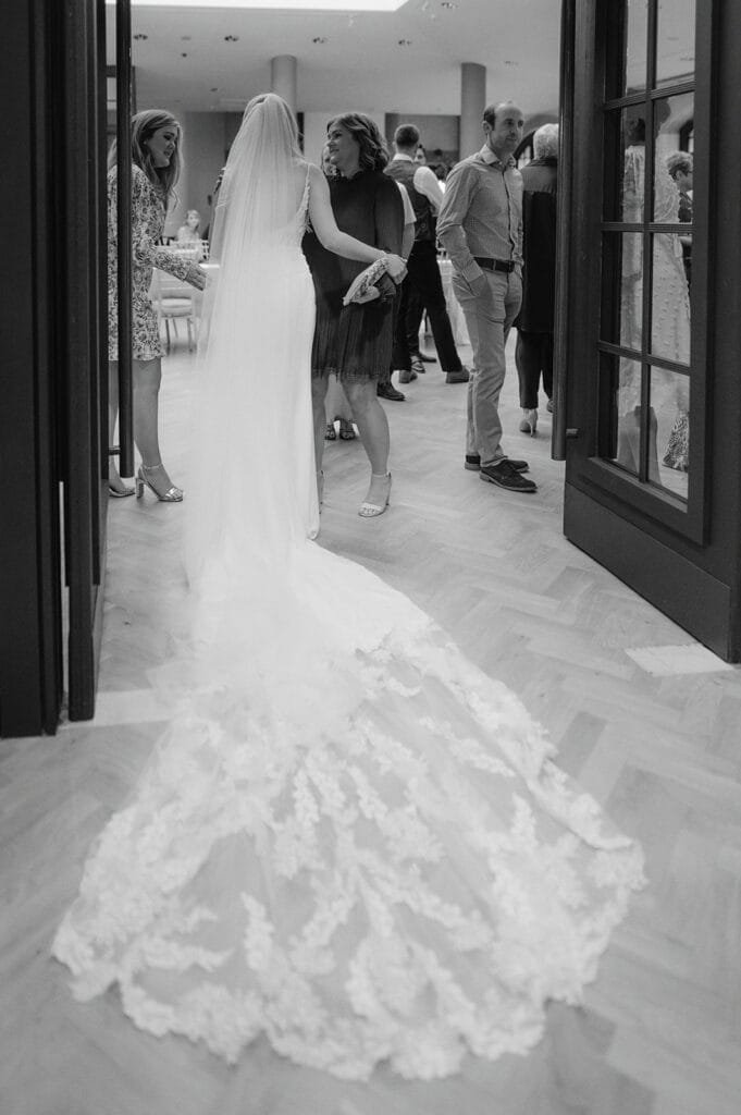 In a timeless black-and-white image, captured by a skilled wedding photographer, a bride in a long lace dress and veil stands gracefully on the wooden floor of The Pumping House. She holds an older womans arm while guests mingle elegantly in the background.