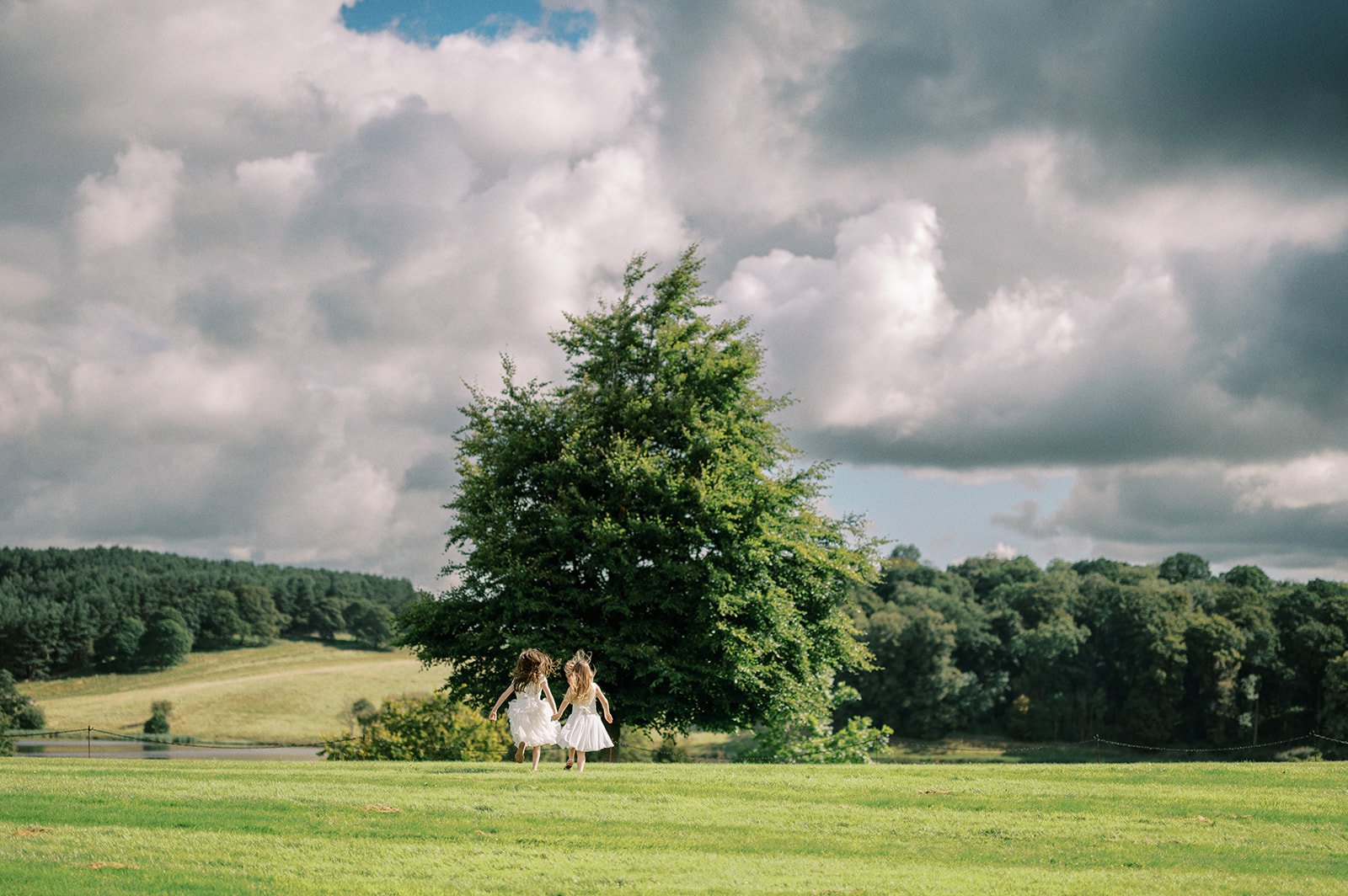 Two children in white dresses run hand in hand across a lush green field towards a large solitary tree, perfect for capturing timeless moments like those of The Coniston Hotel wedding photographer. Dramatic clouds fill the sky, and a forest sits in the background, suggesting a peaceful, rural setting.