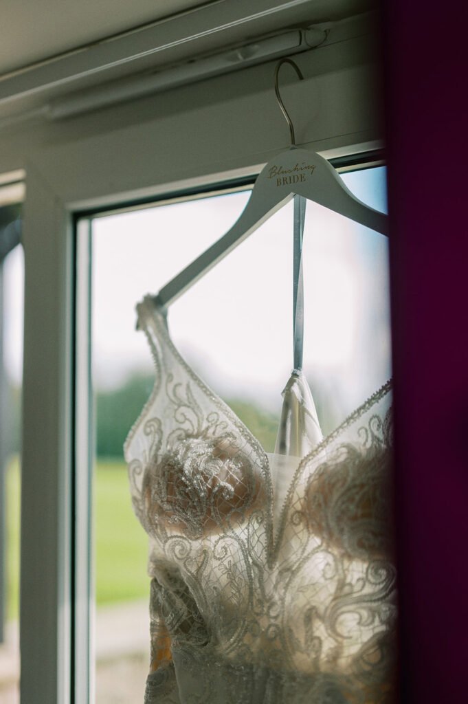 A detailed lace wedding dress is hanging on a white hanger marked BRIDE in cursive. Its displayed by a window at the Coniston Hotel, where natural light highlights the intricate lace patterns. A perfect capture for any discerning wedding photographer.