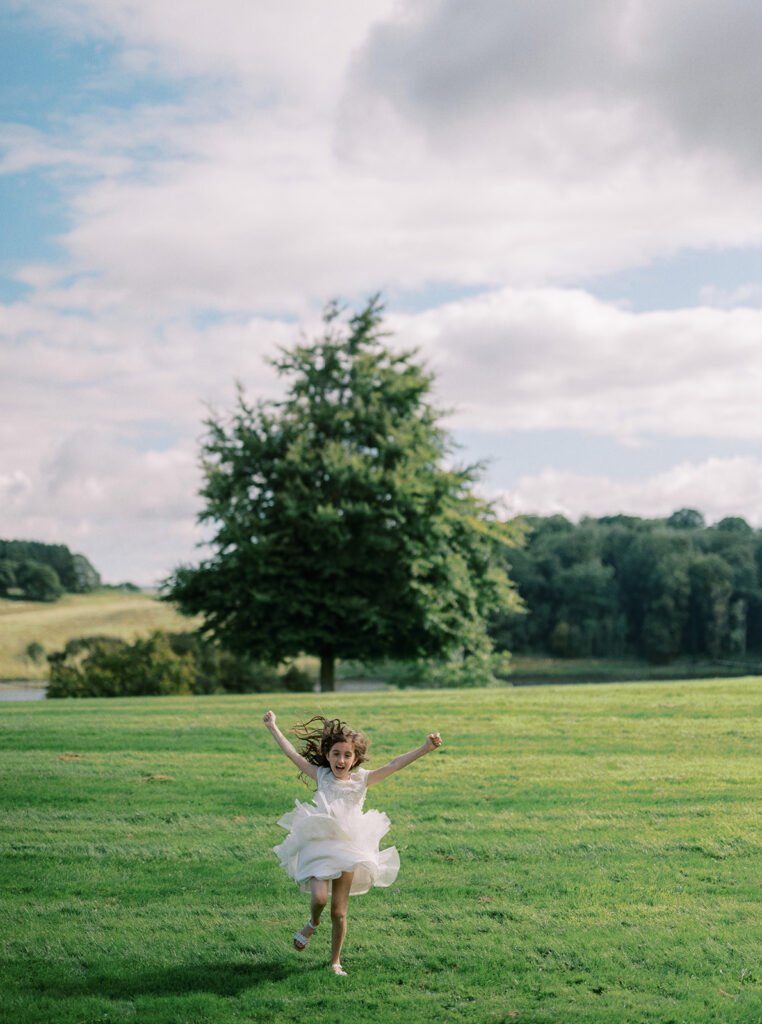 A young girl in a white dress joyfully runs on a grassy field, perfect for capturing as a Coniston Hotel wedding photographer would. Her outstretched arms and flowing hair are set against the backdrop of a large tree, scenic hills, and a cloudy sky.