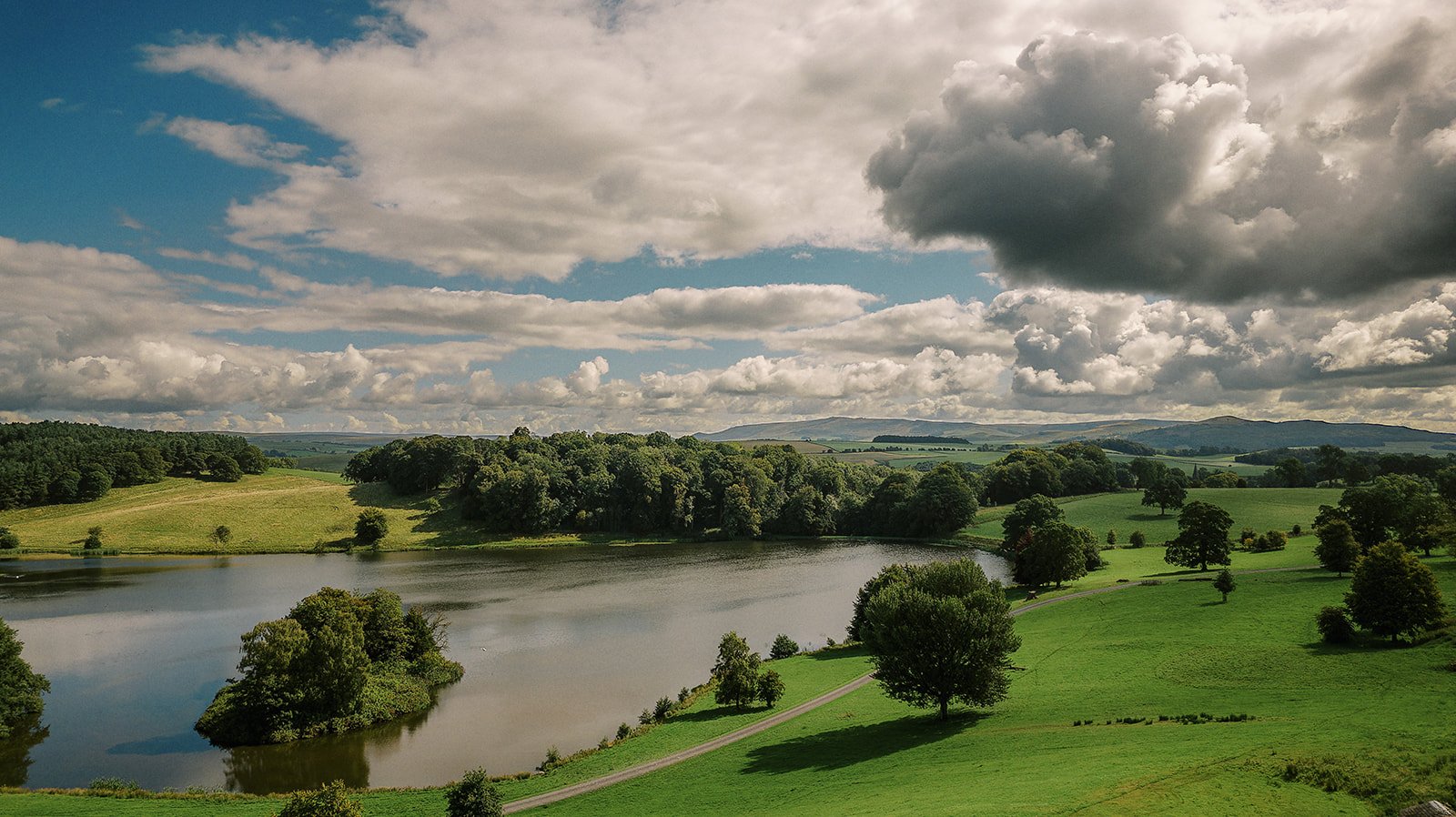 A serene landscape unfolds like a Coniston Hotel wedding photographers dream, featuring a tranquil lake surrounded by lush, green fields and trees under a partly cloudy sky. Rolling hills are visible in the distance, with sunlight casting gentle shadows on the picturesque scene.